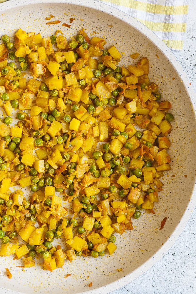 A white frying pan from above with chopped potatoes, peas, shredded carrots turned yellowish due to the seasoning added. 