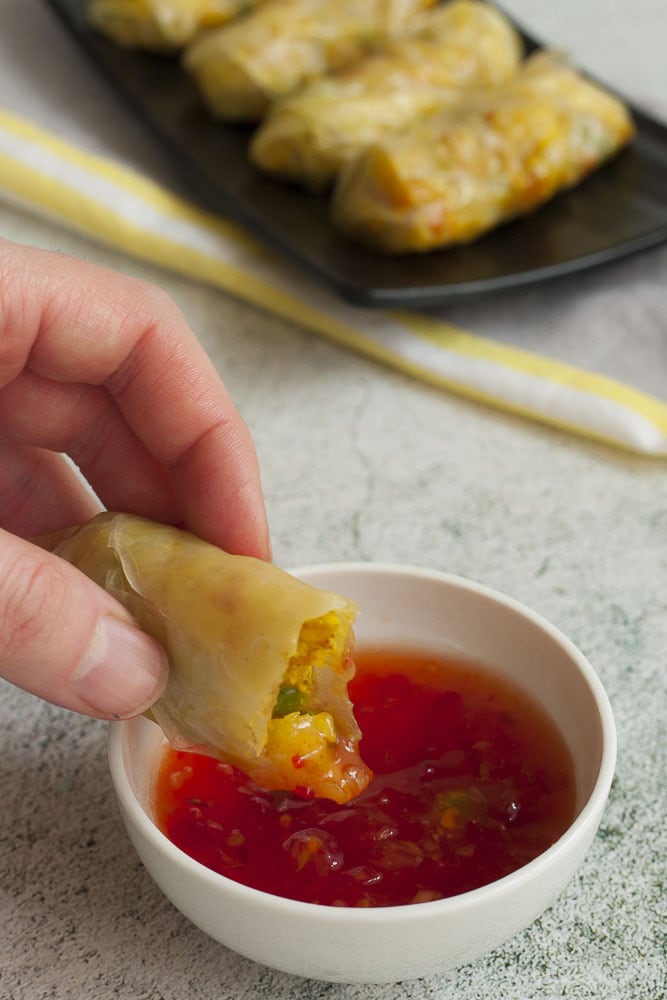 A hand is holding the yellow roll about to dip it into a red sauce in a white bowl.