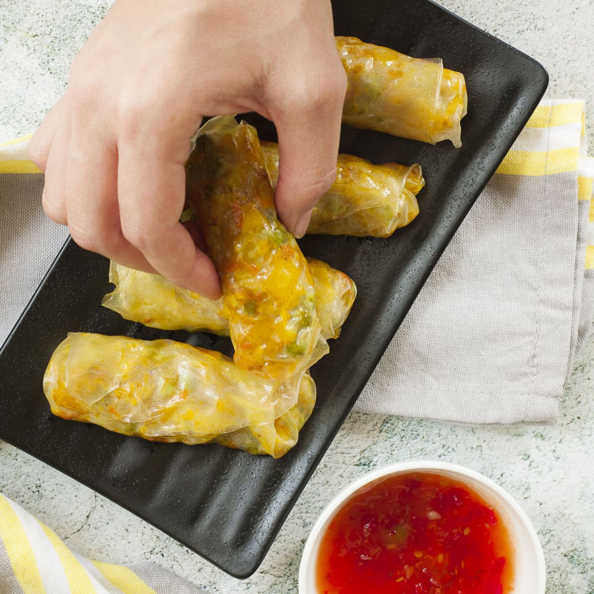 Black plate with 4 yellow rolls. A hand is holding the fifth roll about to dip it into a red sauce in a white bowl.