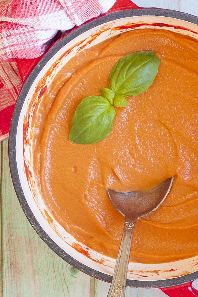 A red Dutch oven full of vegetable pasta sauce topped with fresh basil leaves photographed from above on a blue board. 