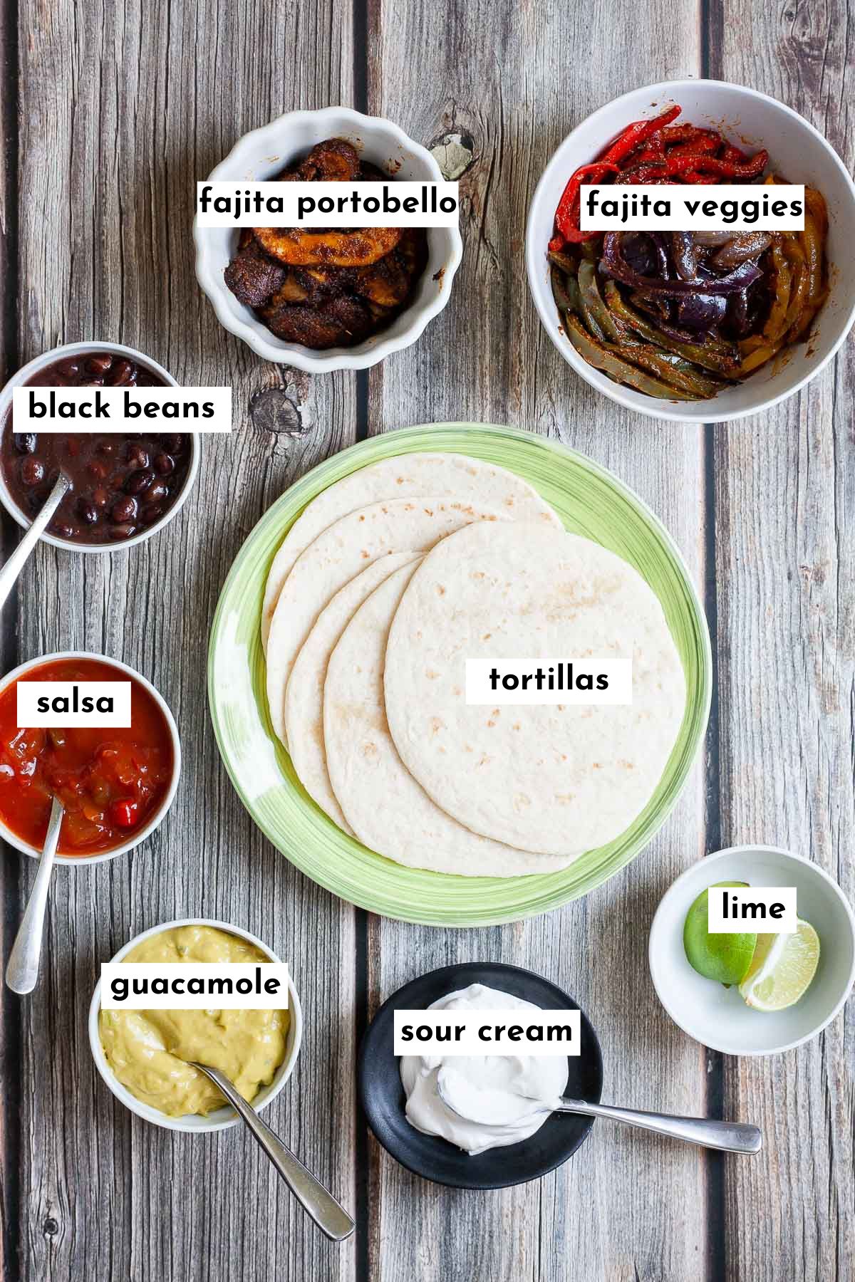 Ingredients to build a mushroom fajitas is in small bowls like guacamole, mushroom slices, veggie slices, black beans, salsa, sour cream, lime and soft tortillas on a green plate