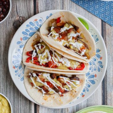 3 soft tortillas folded in half on a white blue plate filled with red pepper slices, purple onion slices and mushroom slices drizzled with white sauce, red salsa and guacamole.