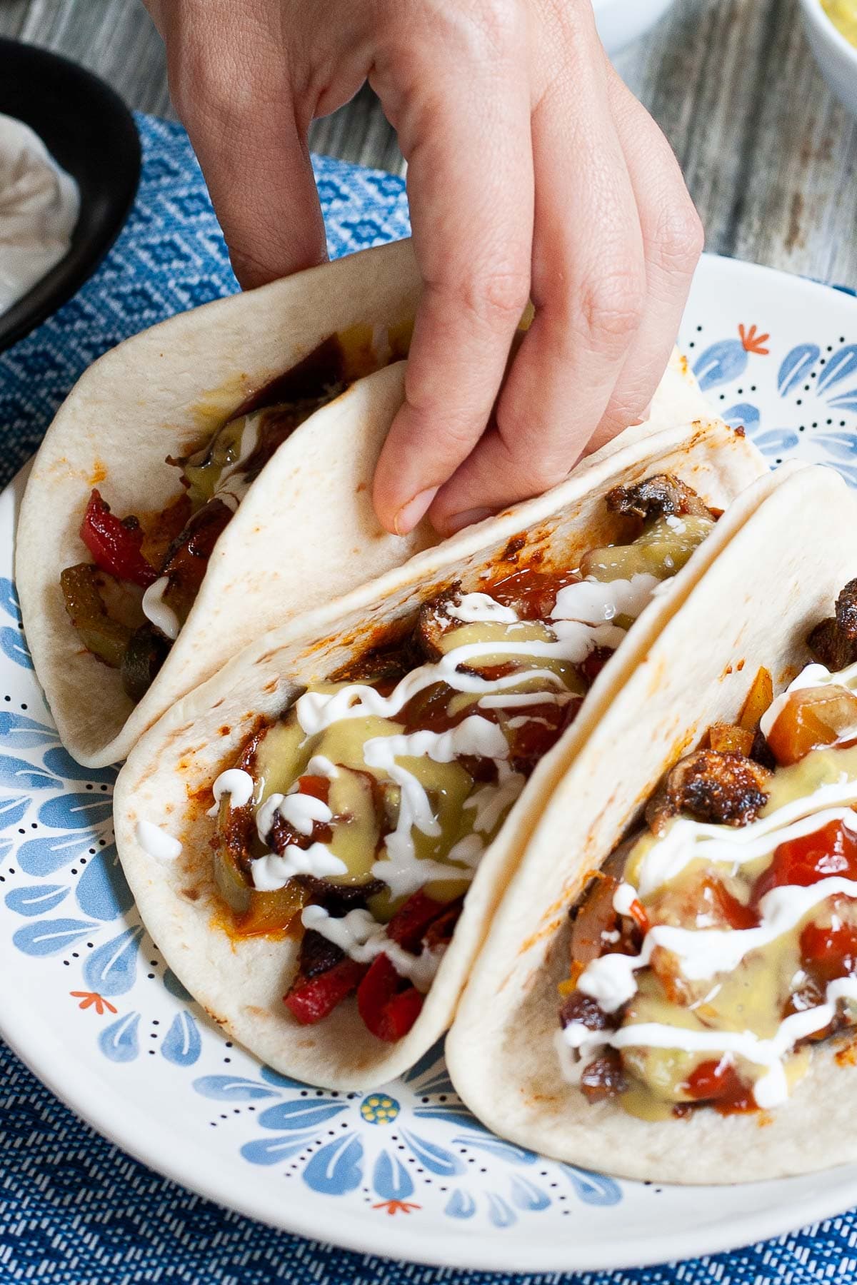 3 soft tortillas folded in half on a white blue plate filled with red pepper slices, purple onion slices and mushroom slices drizzled with white sauce, red salsa and guacamole. A hand is holding one.