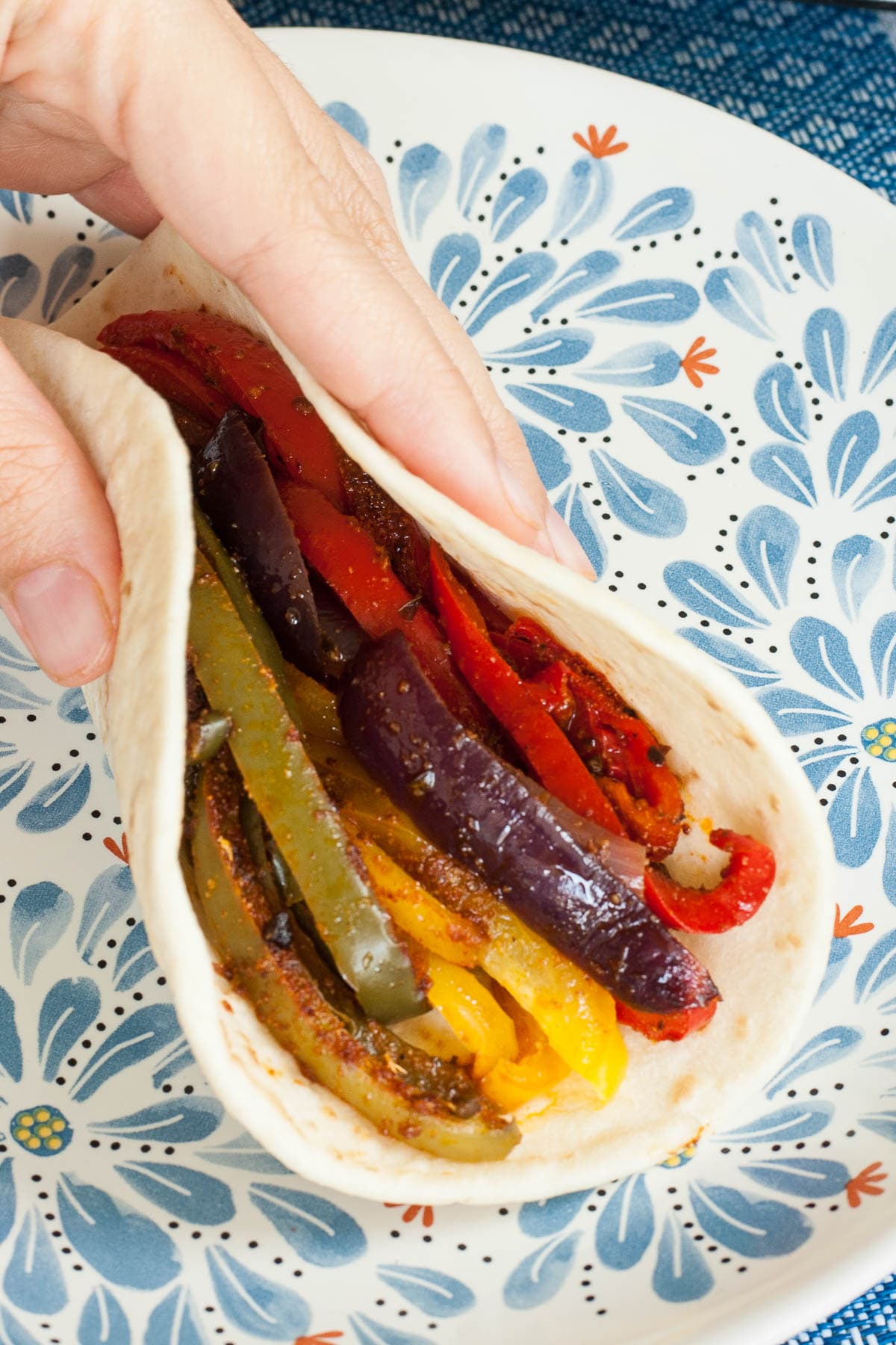 A hand is holding a folded tortilla with red yellow and green bell pepper slices and purple onion slices in front of a blue floral plate