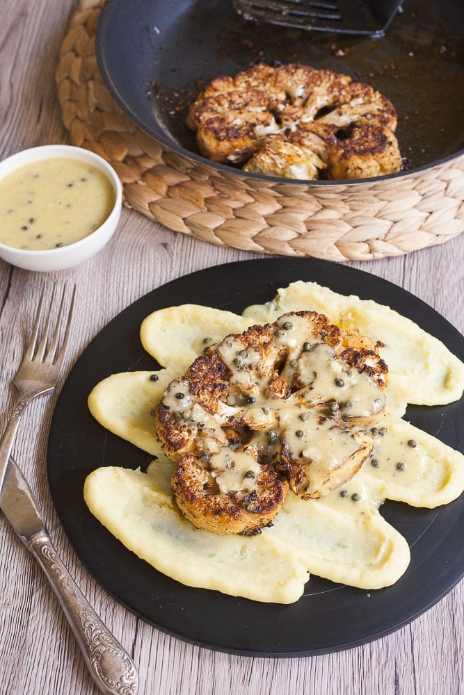 A large slice of brown edged / charred cauliflower topped with a greenish sauce with green peppercorn is placed on top of mashed potatoes. 