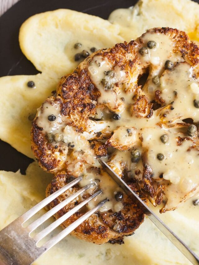 A large slice of cauliflower steak drizzled with a light green sauce with green peppercorns and placed on top of mashed potatoes. A fork and a knife is about to cut in it. 