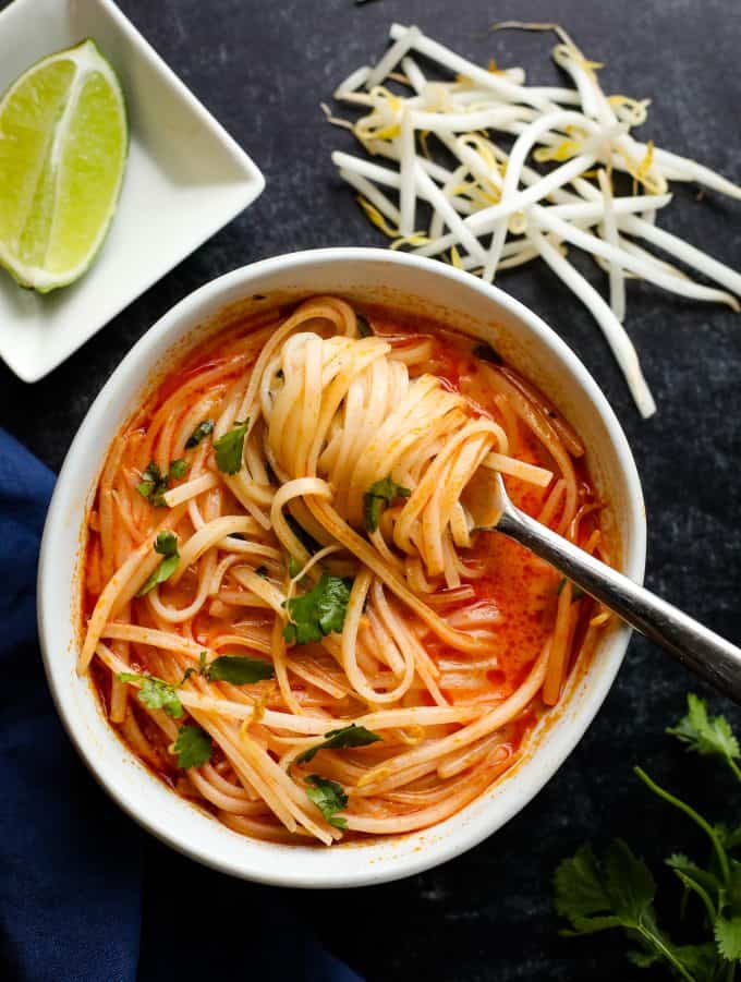 White bowl with red soup, noodles, and chopped green herbs.