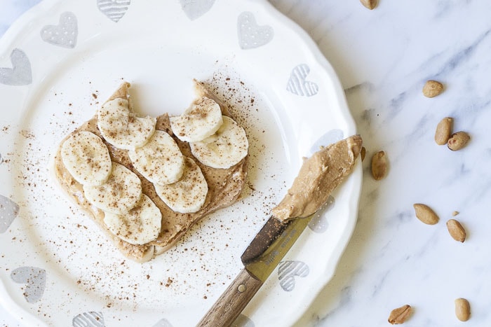 Slice of toast topped with peanut butter, banana slices and cinnamon on a white plate with grey hearts. 