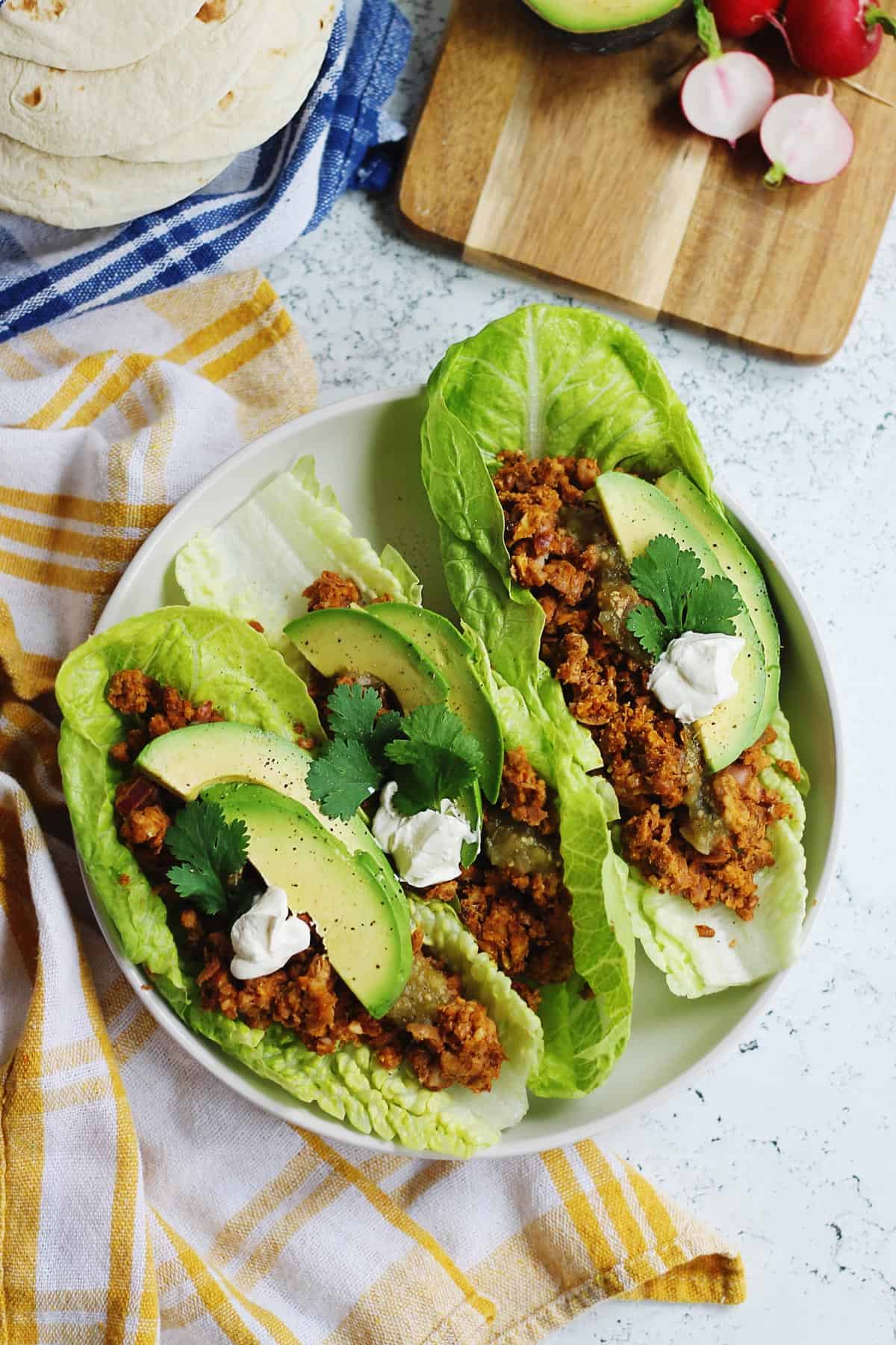 White plate with two lettuce leaves filled with brown crumbles, avocado slices, topped with a white cream and chopped green herbs.