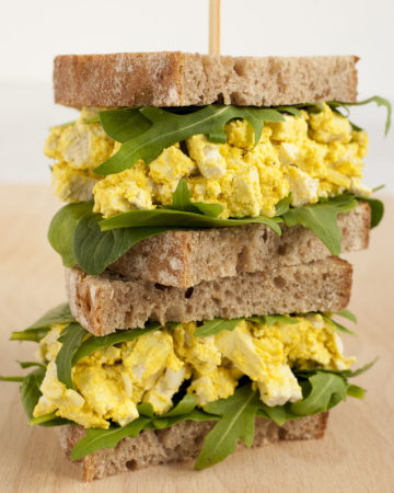 A stack of two sandwiches. Both with brown sliced bread, arugula, and tofu cubes in thick yellow sauce to mimic egg salad.