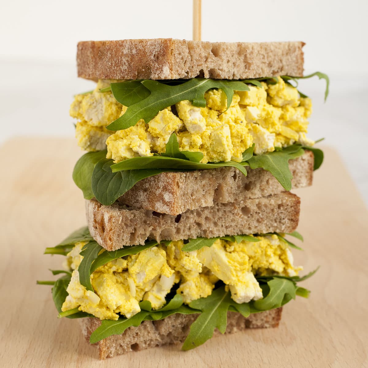 Two vegan egg salad sandwiches on top of each other with arugula and spinach leaves