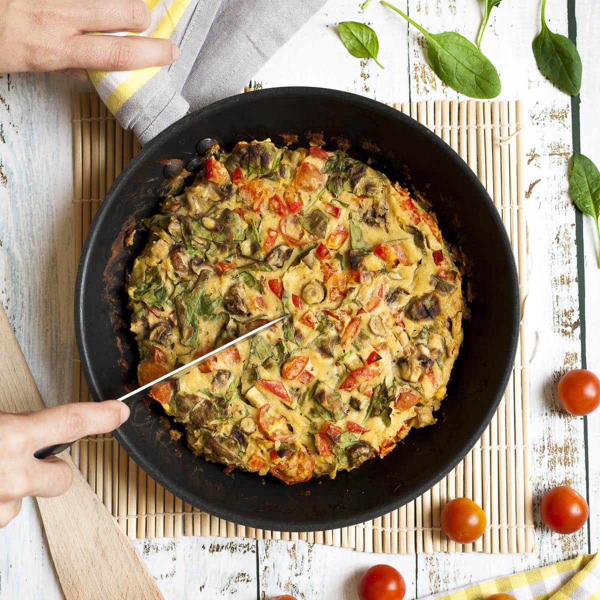 Frying pan from above with a yellow frittata full of veggies like chopped red bell pepper, green spinach leaves, brown mushroom, red tomatoes. A hand is holding a knife and about to slice in it. 