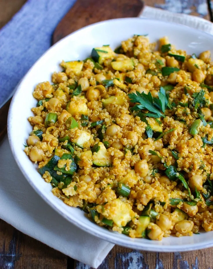 White bowl with quinoa, zucchini, chickpeas in a colored yellow-brown by some spices. Sprinkled with freshly chopped herbs.