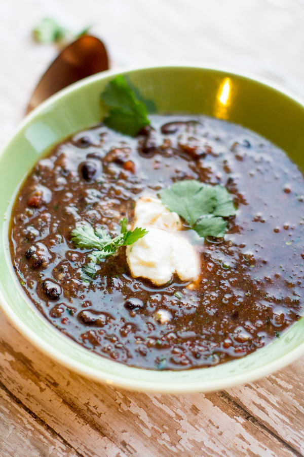 Green bowl with dark brown soup and black bean pieces. Topped with a white cream and chopped green herbs.