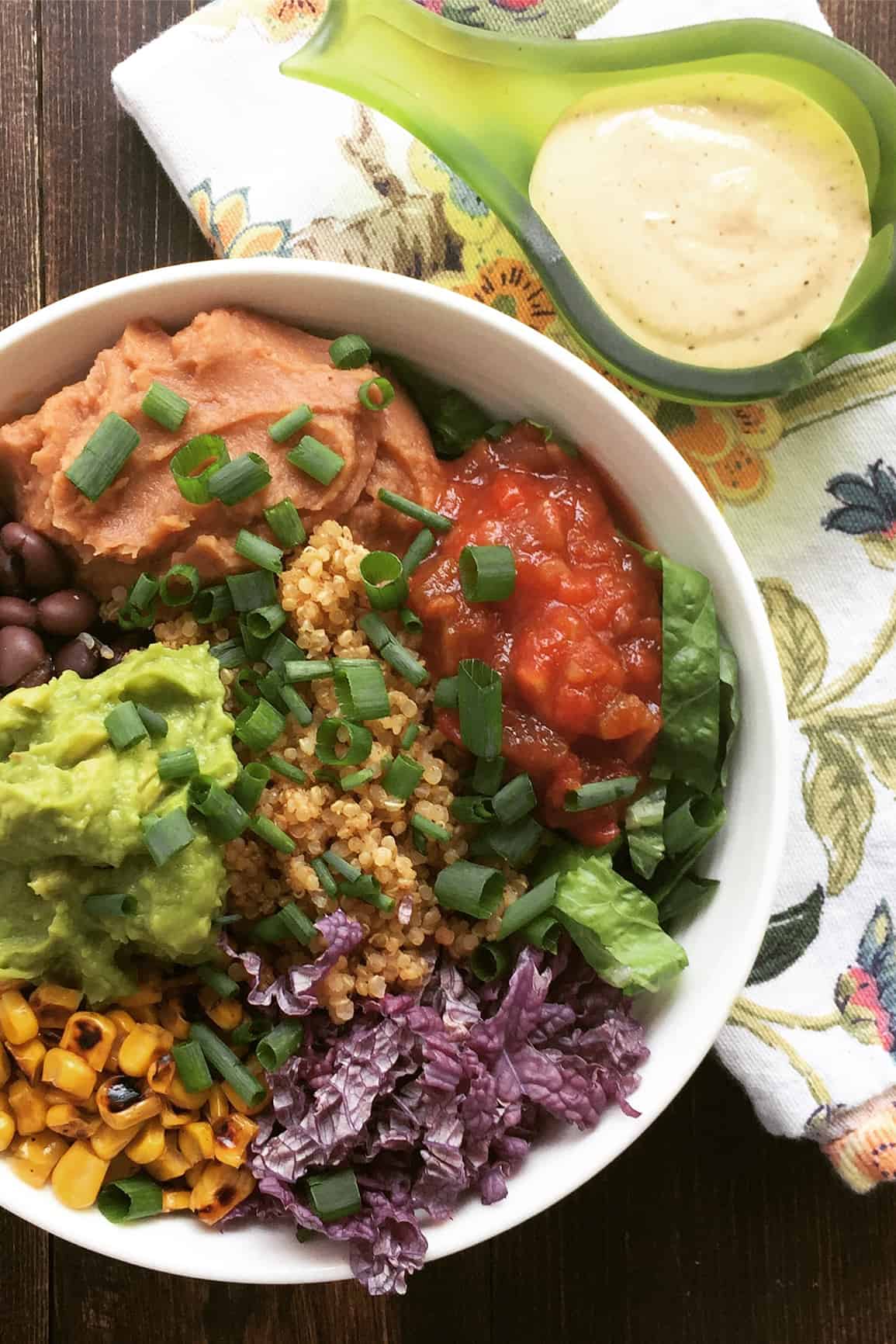 White bowl with roasted corn, avocado mash, black beans, lettuce strips, chopped purple onion, quinoa and red and orange sauces.