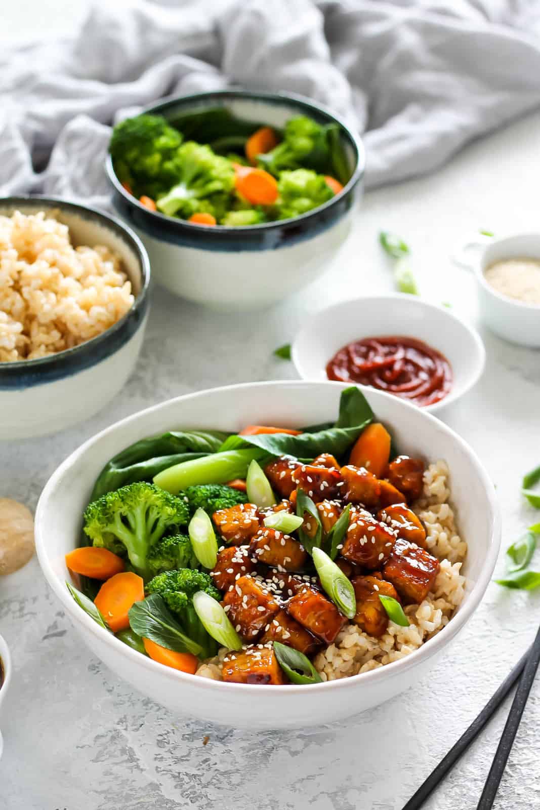 White bowl with brown rice, sticky brown tempeh cubes, carrots slices, broccoli florets and bok choy.