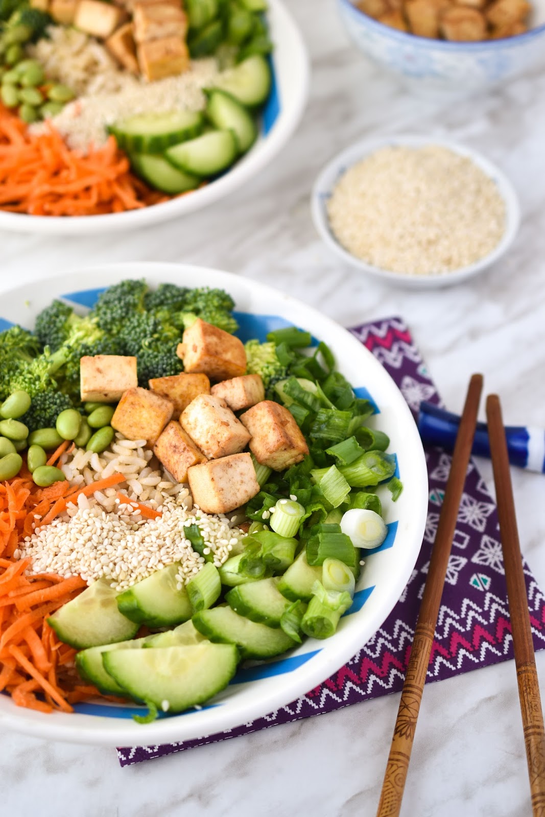 White bowl with cucumber slices, matchstick carrots, scallion, broccoli, crispy tofu cubes, edamame beans, and sesame seeds.