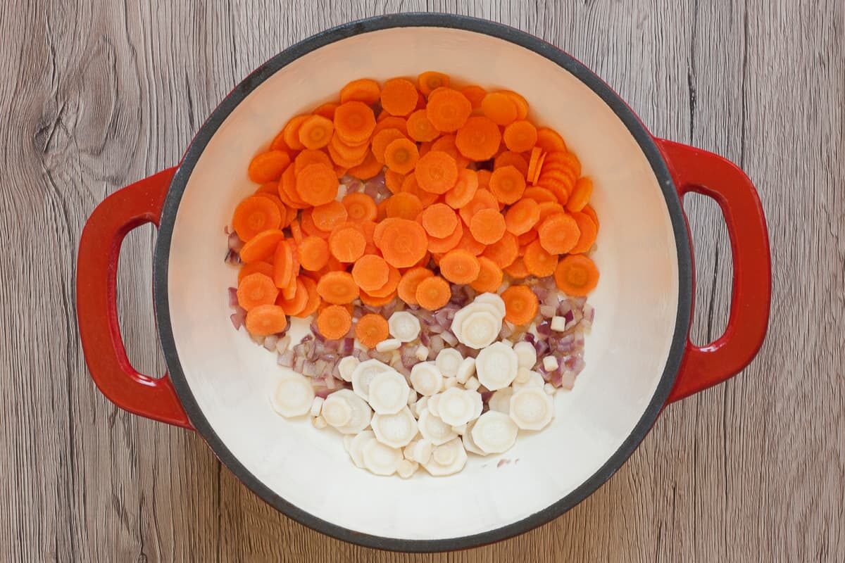 White red enameled Dutch oven with chopped red onion, carrot and parsnip slices.