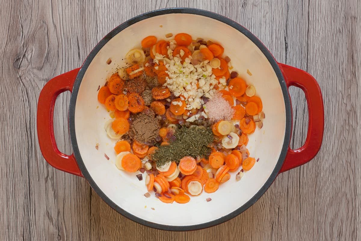 White red enameled Dutch oven with chopped red onion, carrot and parsnip slices and small heaps of different herbs.