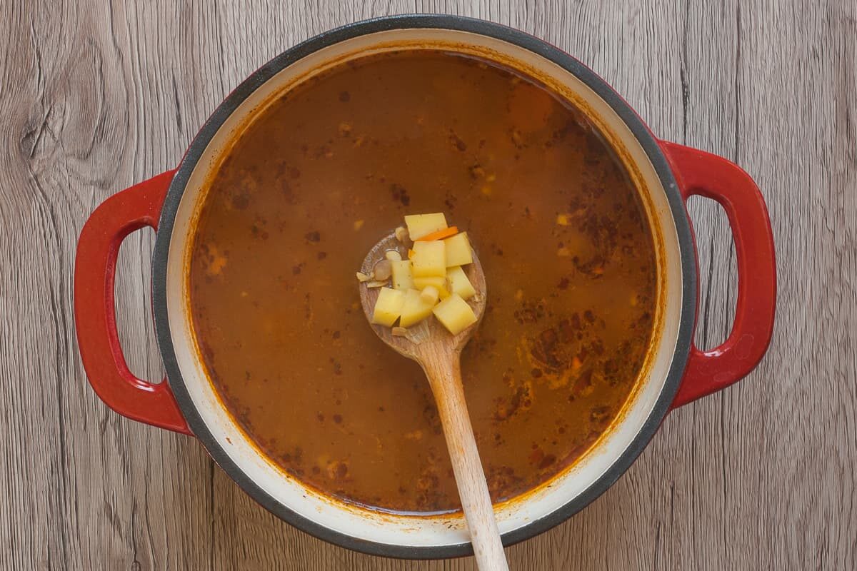 White red enameled Dutch oven with brown soup. A wooden spoon is taking some potato pieces from the soup.