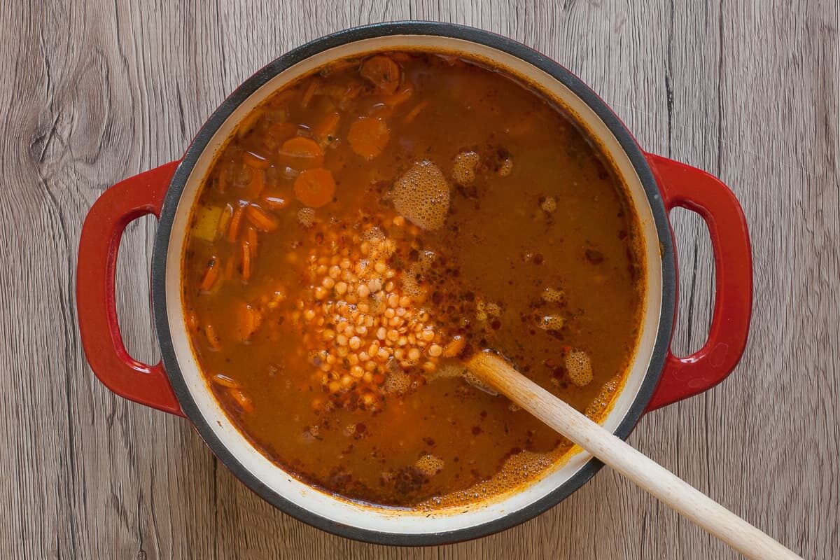 White red enameled Dutch oven with brown soup. A wooden spoon is taking red lentils from the soup.