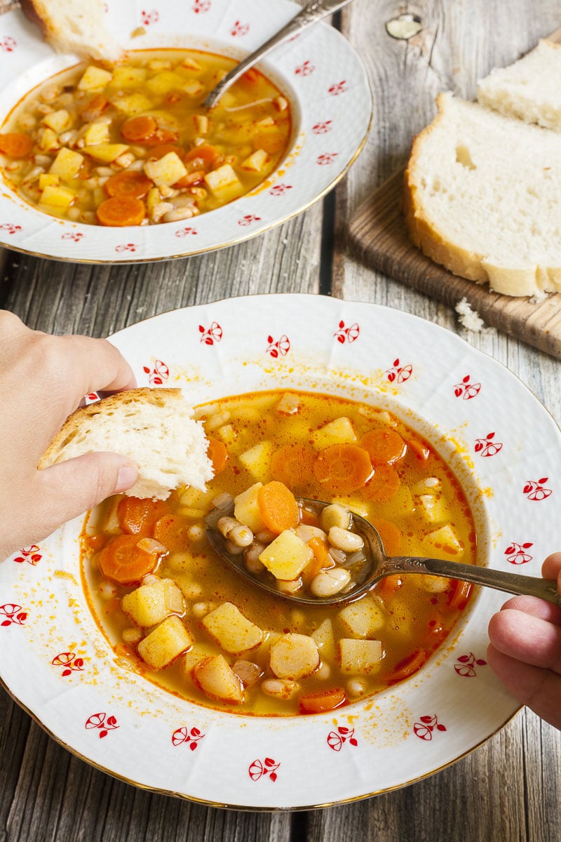 Soup with chopped potatoes, carrots and white beans in 2 white bowls with Hungarian flower patterns. A hand is taking a spoonful and dunks a bread slice.