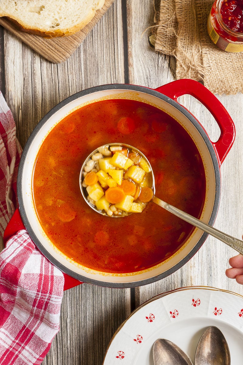A large red white pot with soup. A hand is holding a ladle and taking chopped potatoes, carrots and white beans