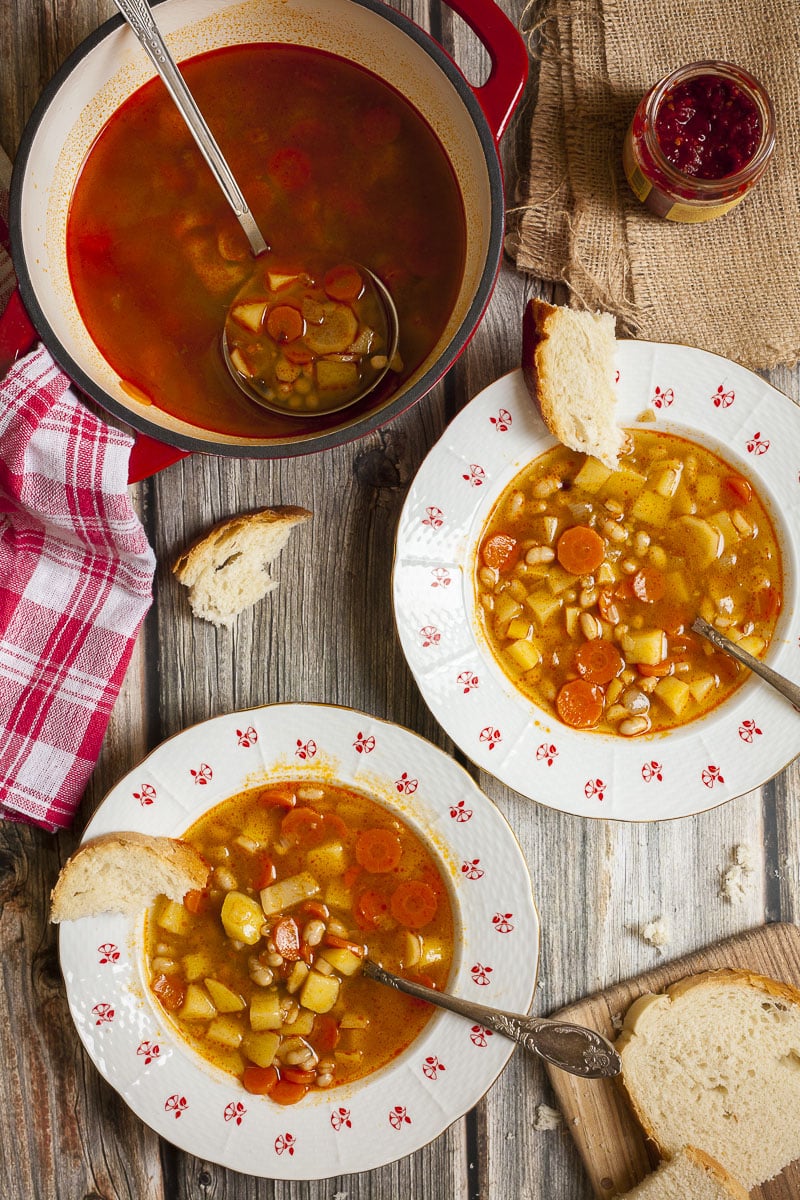 Two plates of soups with chopped potatoes, carrots and white beans. Slices of bread and spoons are next it as well as a large white and red pot with ladle. 