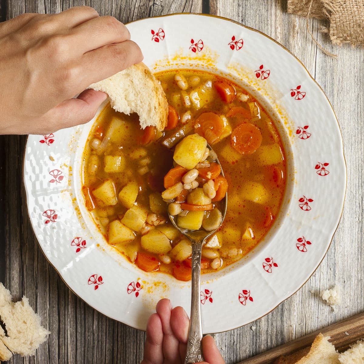 Soup with chopped potatoes, carrots and white beans in a white bowl with Hungarian flower patterns. A hand is taking a spoonful and dunks a bread slice