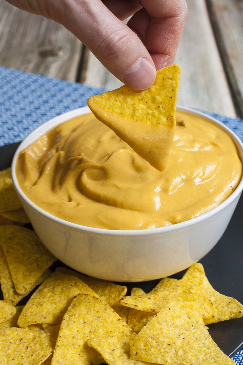 White bowl of cheese sauce and a hands dips a nachos chips