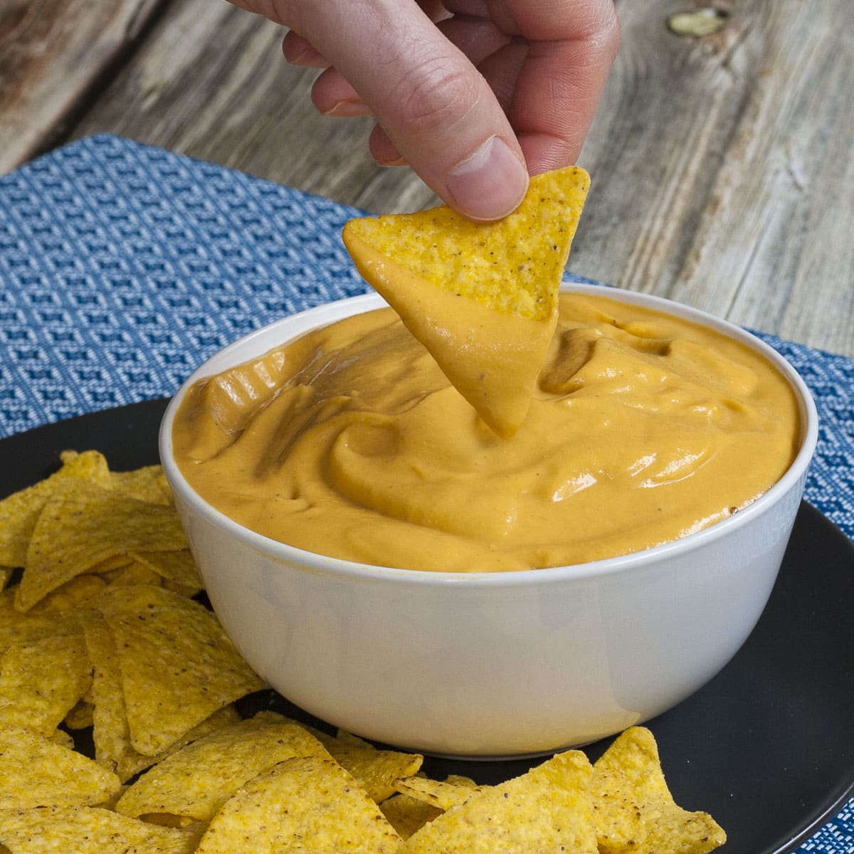 White bowl of vegan nacho cheese sauce and a hand dips a tortilla in it