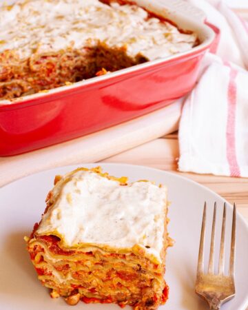 A slice of layered lasagna is served on a plate. A red casserole dish with the remaining lasagna is right behind it.