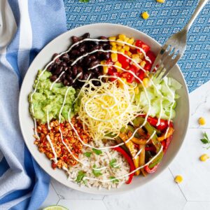 A large white plate in the middle with colorful ingredients like black beans, corn, shredded cheese, bell pepper strips, rice, shredded lettuce, avocado.