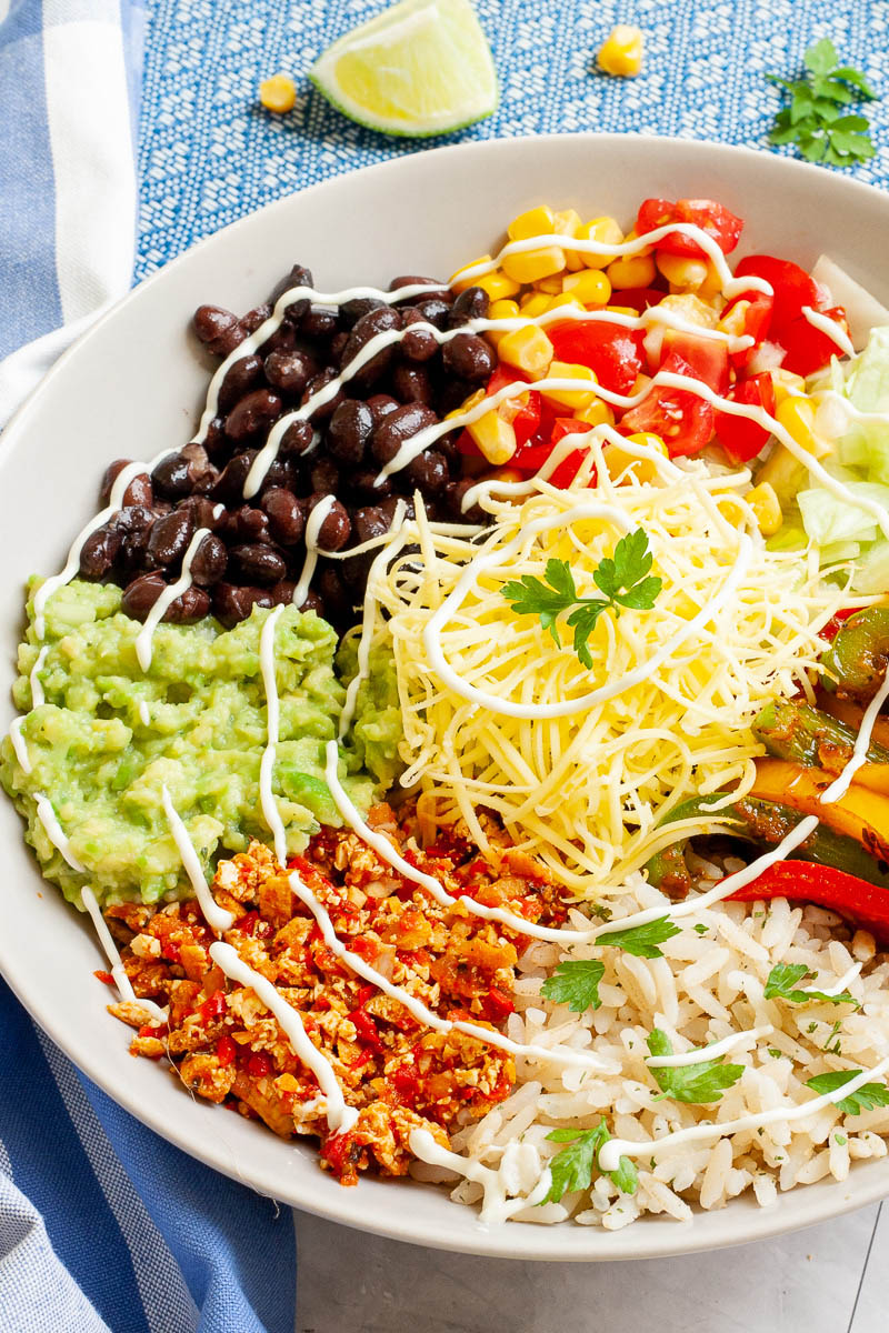 A large white plate in the middle with colorful ingredients like black beans, corn, shredded cheese, bell pepper strips, rice, shredded lettuce, avocado.