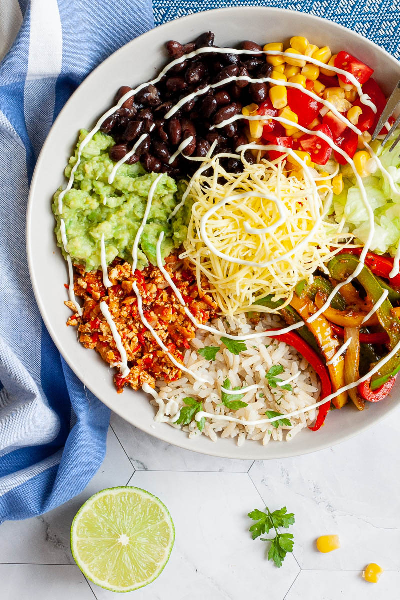 A large white plate in the middle with colorful ingredients like black beans, corn, shredded cheese, bell pepper strips, rice, shredded lettuce, avocado. Blue white table clothes with scattered corn and lime wedges.