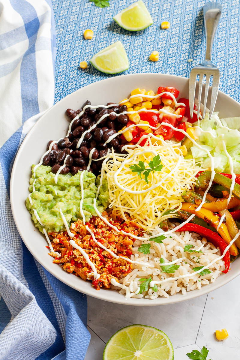A large white plate in the middle with colorful ingredients like black beans, corn, shredded cheese, bell pepper strips, rice, shredded lettuce, avocado. Blue white table clothes with scattered corn and lime wedges.