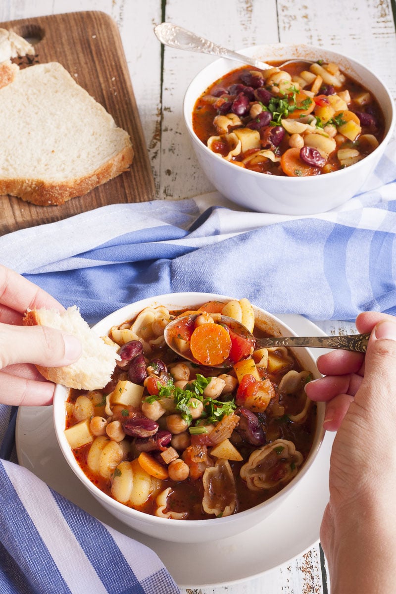 A hand is dunking a bread and taking a spoonful from a red soup served in a white bowl that is full of red kidney beans, chickpeas, potatoes, carrots and shell pasta. It is topped with fresh parsley. 