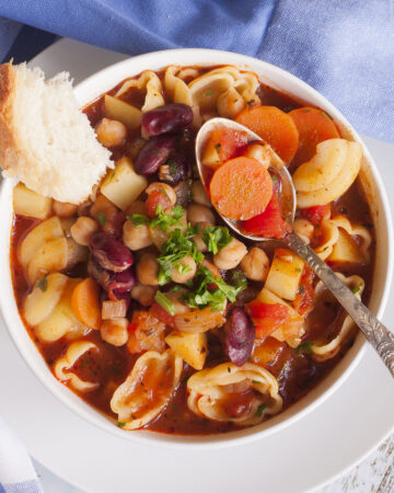 White bowl with a red soup full of red kidney beans, chickpeas, potatoes, carrots and shell pasta. It is topped with fresh parsley. A spoon is taking a carrot.