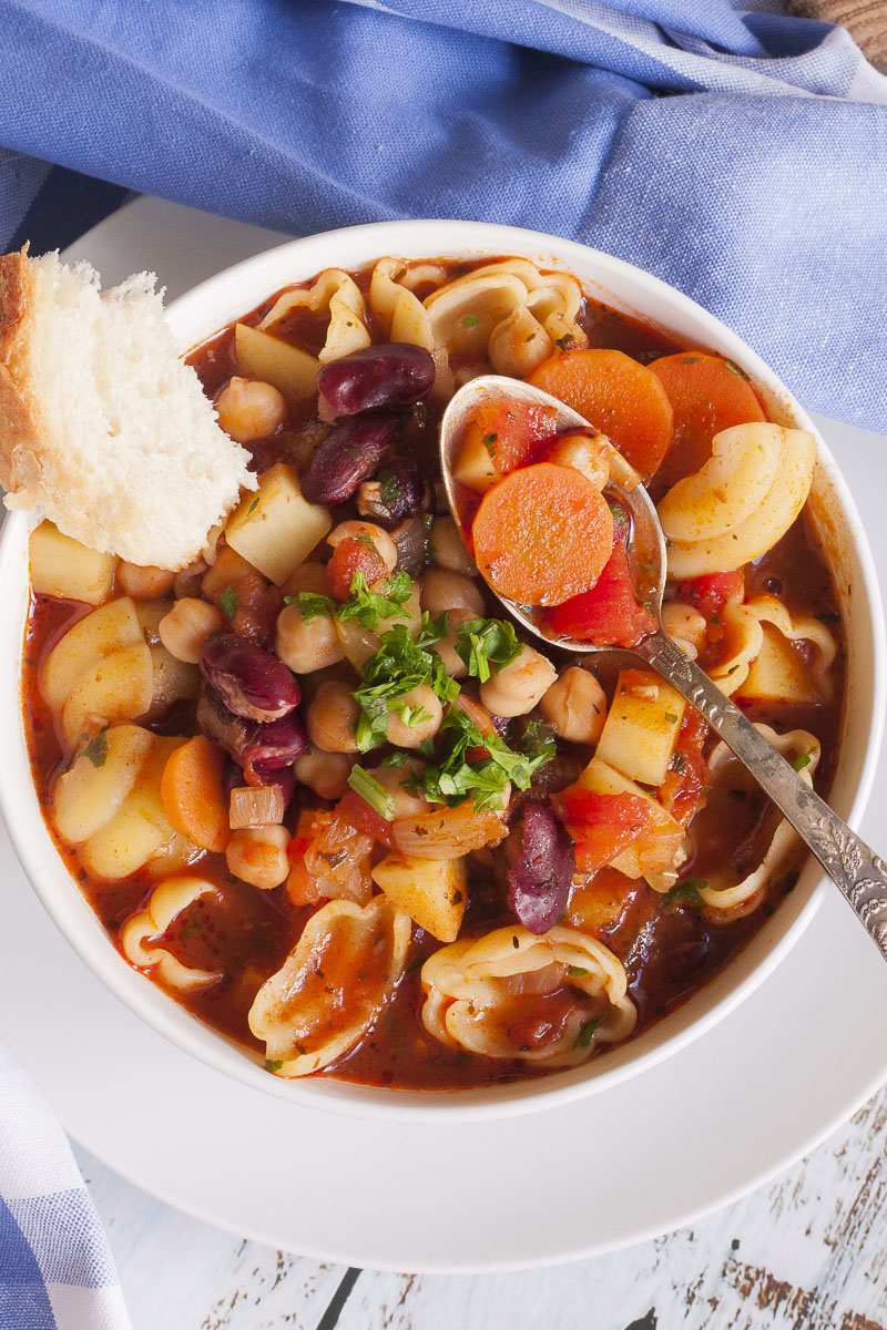 White bowl with a red soup full of red kidney beans, chickpeas, potatoes, carrots and shell pasta. It is topped with fresh parsley. A spoon is taking a carrot.