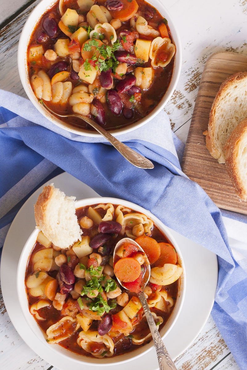 2 white bowls with a red soup full of red kidney beans, chickpeas, potatoes, carrots and shell pasta. It is topped with fresh parsley. A spoon is taking a carrot.