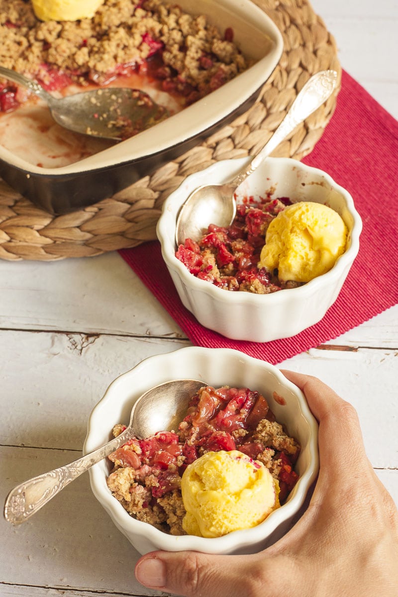 2 small white bowls with red fruit and brown crisp topping with vanilla ice cream. The baking dish with the remaining portions is in the background. 