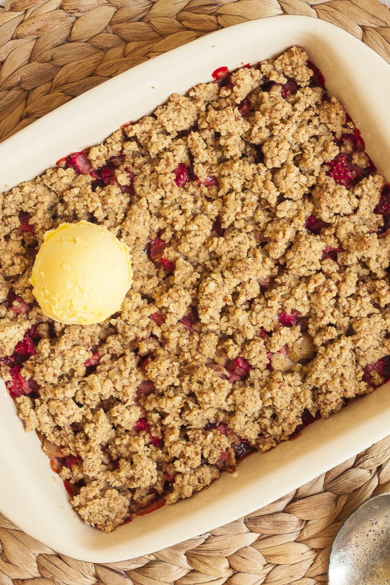 A white rectangle baking dish from above with brown crumb-like topping and red sauces peaking out of it. It is topped with one scoop of vanilla ice cream