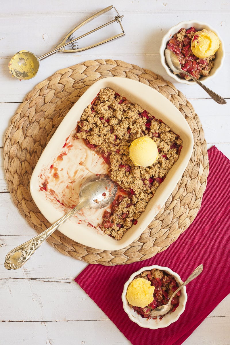 A white rectangle baking dish from above with brown crumb-like topping and red sauces peaking out of it. It is topped with one scoop of vanilla ice cream. 2 small white bowl is next to it with red fruity sauce and brown crisp and vanilla ice cream