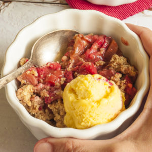 A hand is holding a small white bowl with red fruit, brown crisp topping and vanilla ice cream.