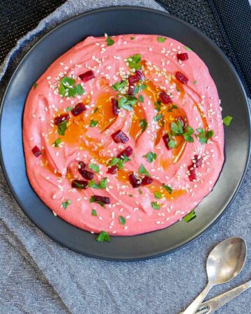Pink hummus on a black plate, sprinkled with freshly chopped parsley, sesame seeds and small roasted beet pieces.