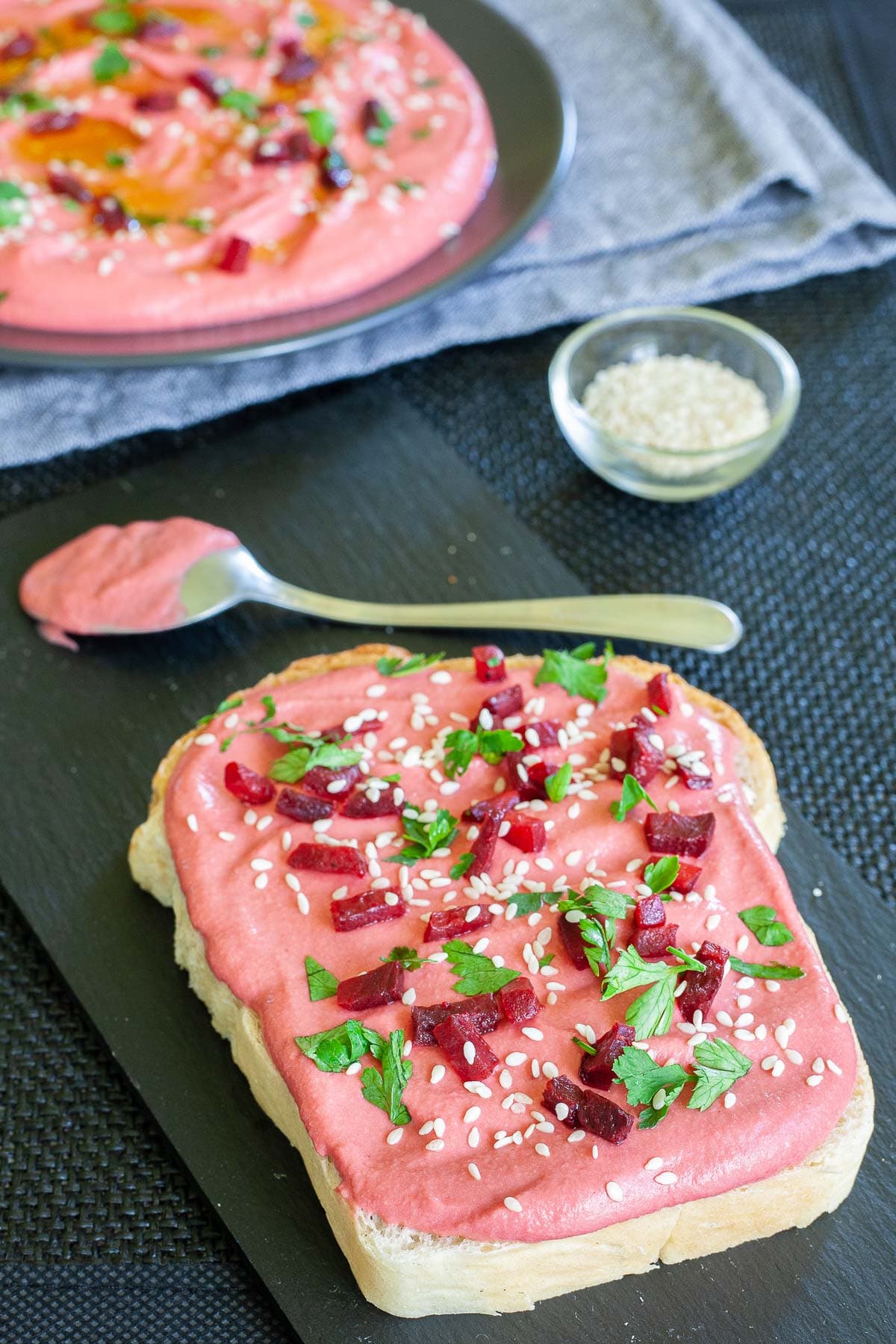 A slice of bread with pink hummus, sprinkled with freshly chopped parsley, sesame seeds and small roasted beet pieces