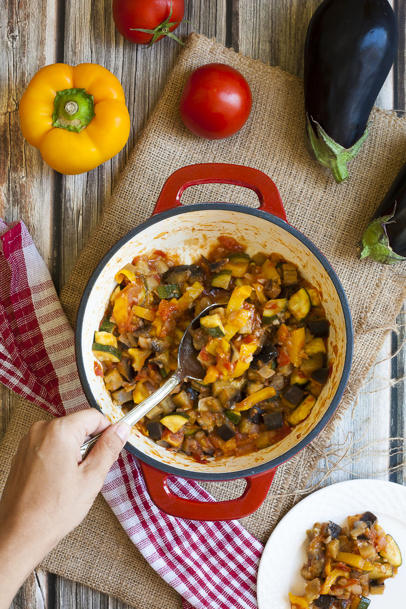 A red pot from above with veggie chunks of all colors: red, yellow, green, orange, brown. There is a serving spoon taking a portion from the middle. 