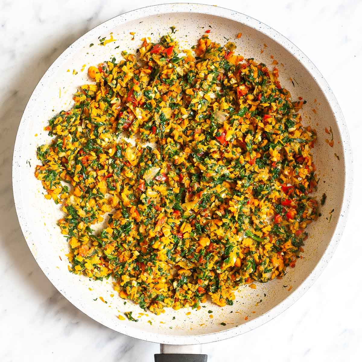 White frying pan with finely chopped spinach, carrots, onion, red pepper, and garlic pieces.