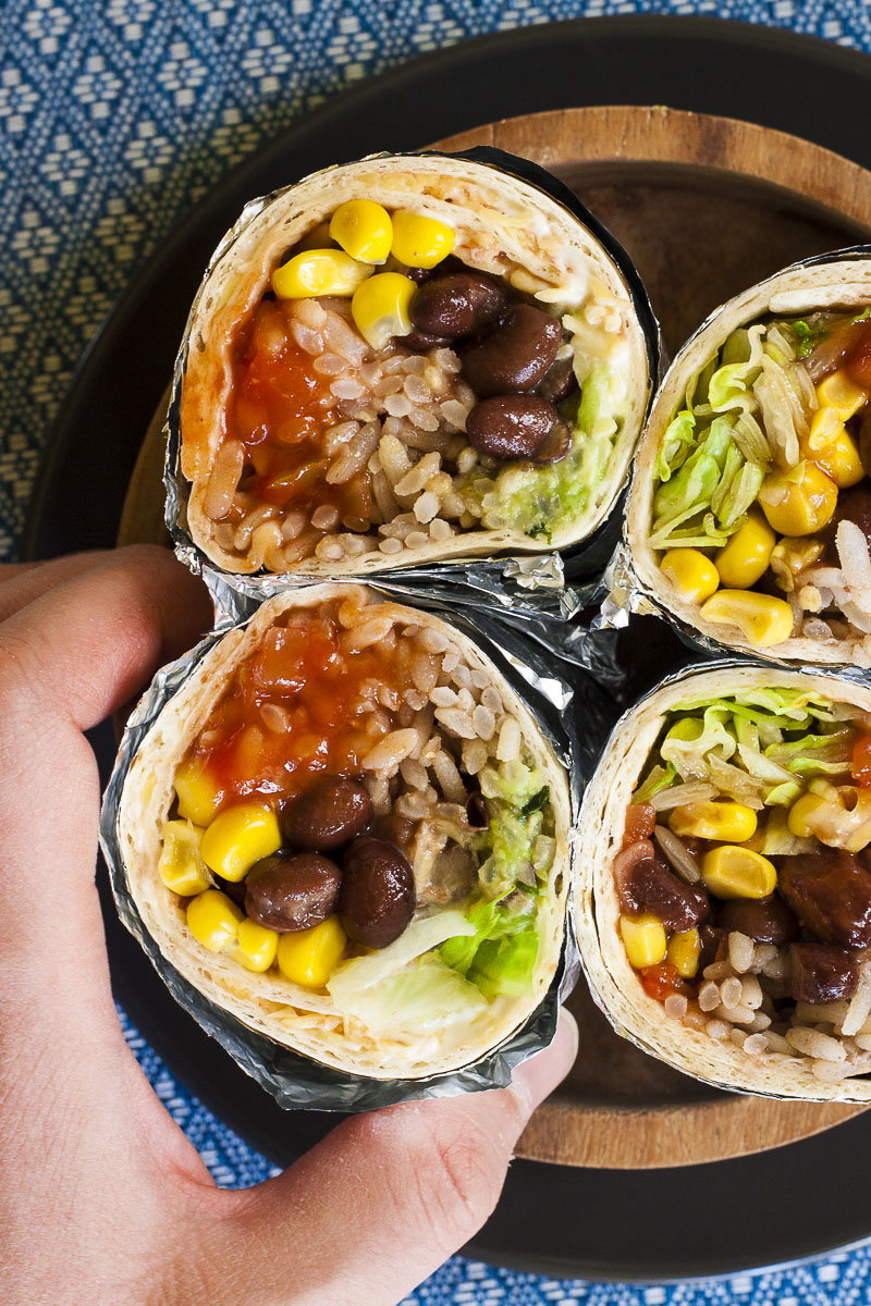 A hand is holding one of the four rainbow coloured round-shaped rolls from above. Inside red, yellow, brown, green and white pieces.