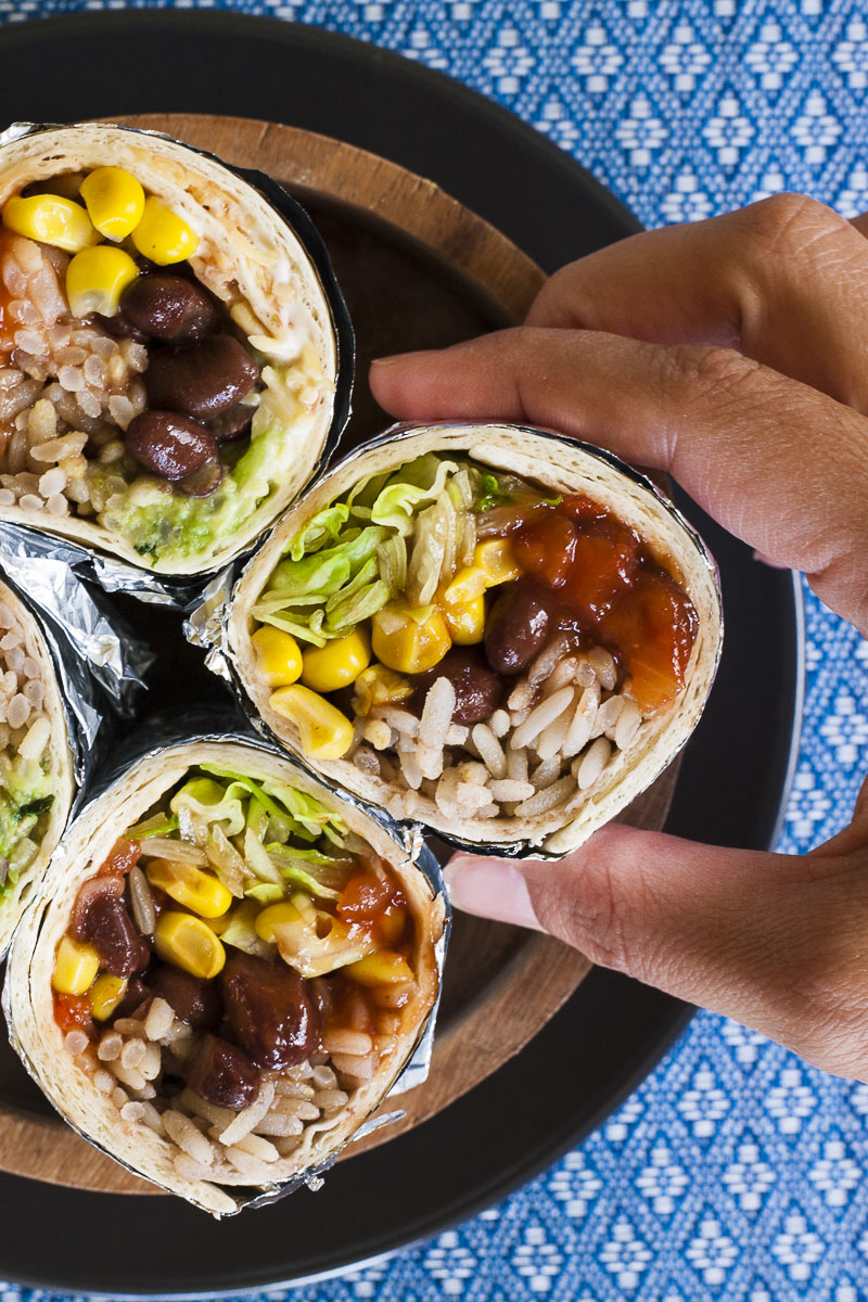 A hand is holding one of the four rainbow coloured round-shaped rolls from above. Inside red, yellow, brown, green and white pieces.